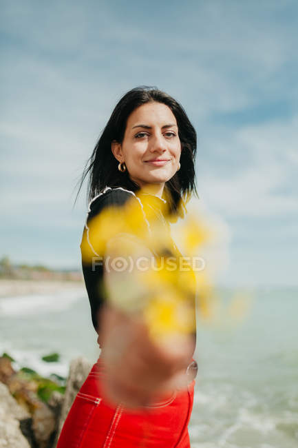 Sorrindo jovem mulher dando um monte de flores amarelas, enquanto em pé na praia no dia ensolarado — Fotografia de Stock