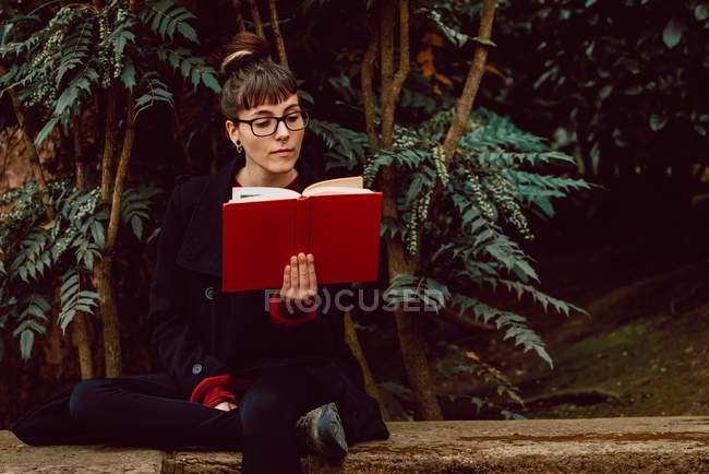 Giovane donna elegante in occhiali libro di lettura e seduta sulla panchina in giardino della città — Foto stock