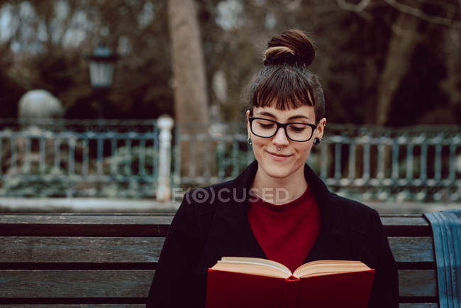 Young elegant woman in eyeglasses reading book and sitting on bench in city garden — Stock Photo