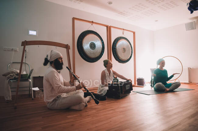 Mulher sentada em lótus posar perto de músicos tocando em instrumentos étnicos perto gongos no quarto — Fotografia de Stock