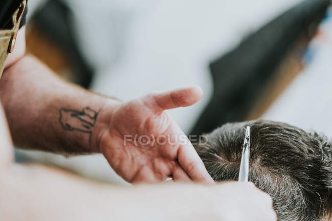 Imagem cortada de barbeiro com tesoura cortando cabelo de macho sentado na barbearia em fundo borrado — Fotografia de Stock