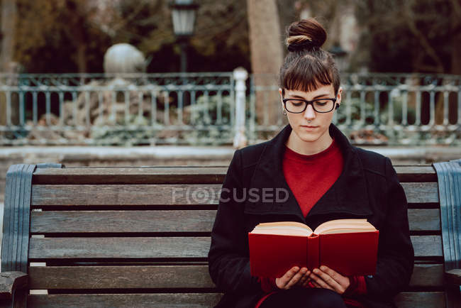 Giovane donna elegante attraente in occhiali da vista lettura libro e seduto sulla panchina in giardino della città — Foto stock