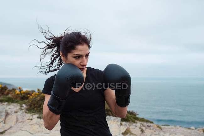 Mulher em luvas de boxe treinando no penhasco contra o mar e o céu — Fotografia de Stock