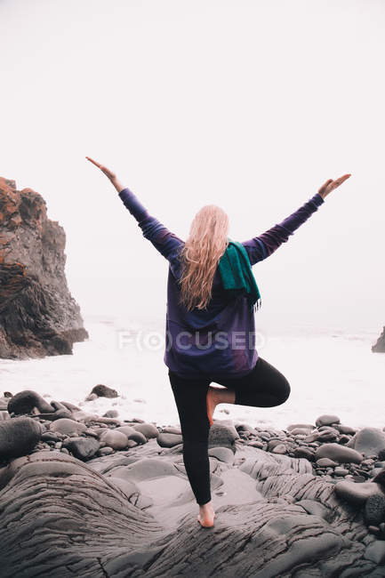 Young woman with upped hands meditating and standing on one leg on rocks on sea coast — Stock Photo