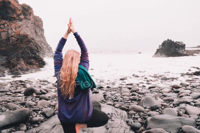 Jovem mulher com as mãos levantadas meditando e de pé em uma perna em rochas na costa do mar — Fotografia de Stock