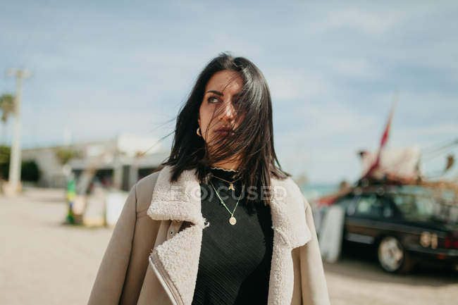Thoughtful young woman in trendy outfit with jacket standing on blurred background of beach parking — Stock Photo