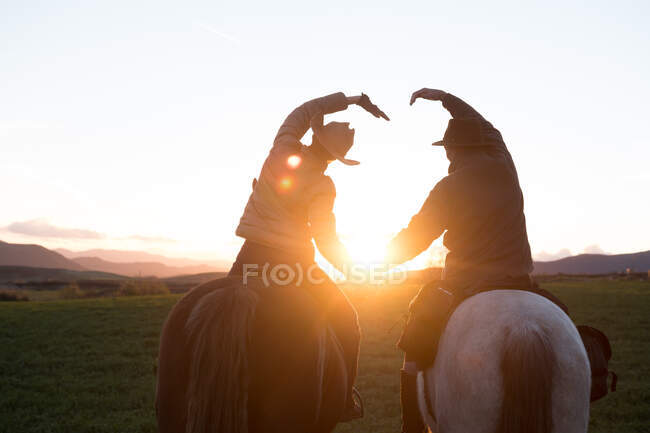 Vue arrière de l'homme et de la femme chevauchant des chevaux et faisant forme de coeur avec les mains contre le coucher du soleil ciel sur ranch — Photo de stock