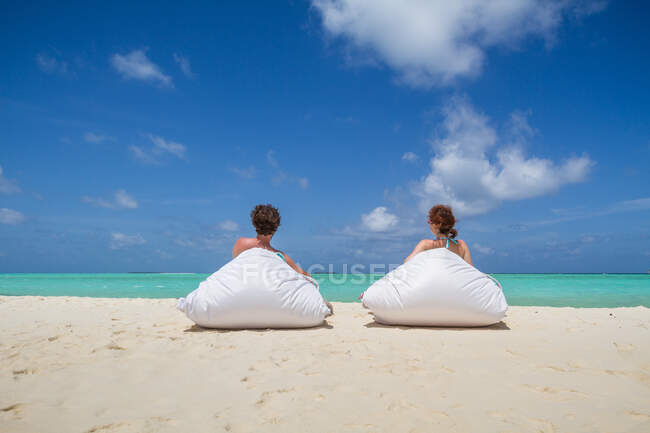 Обратный вид на мужчину и женщину, лежащих на мягких бобовых мешках на песчаном берегу возле удивительного моря против облачного неба в солнечный день на Мальдивах — стоковое фото