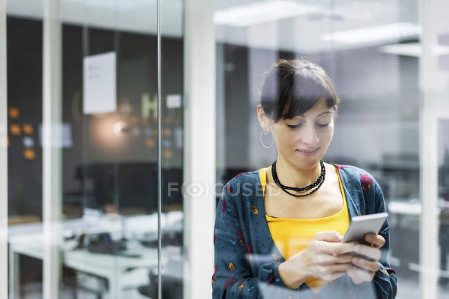 Gestionnaire femme souriante en utilisant smartphone tout en se tenant près du mur de verre dans le bureau moderne — Photo de stock