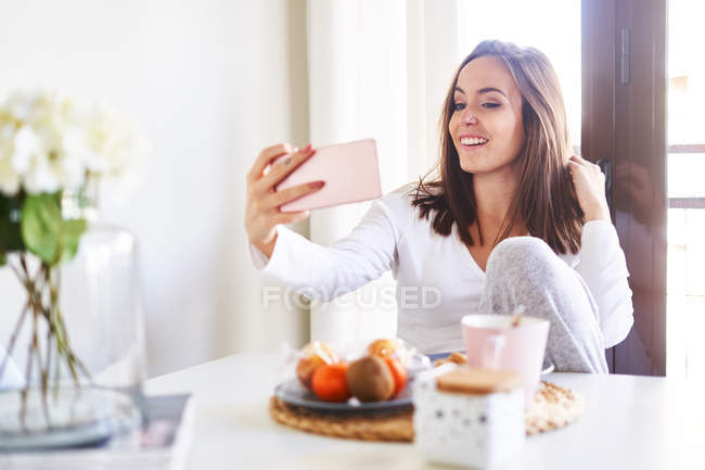 Jovem mulher feliz tomando selfie com telefone celular enquanto toma café da manhã perto da janela em casa — Fotografia de Stock