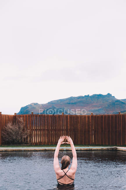 Vue arrière de la jeune femme reposant dans l'eau de la piscine contre une clôture en bois dans la nature avec montagne sur fond — Photo de stock