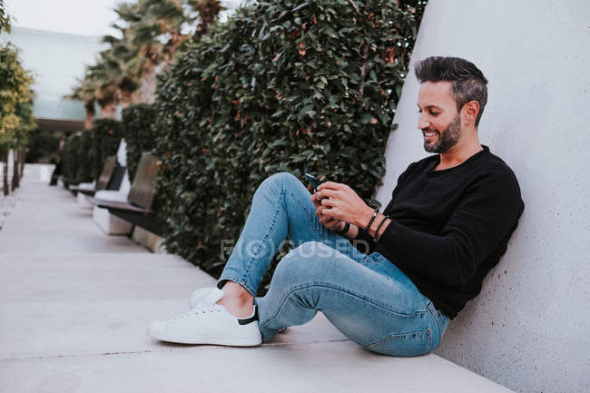 Bel élégant homme heureux en tenue décontractée à l'aide d'un téléphone portable et assis près du mur gris — Photo de stock