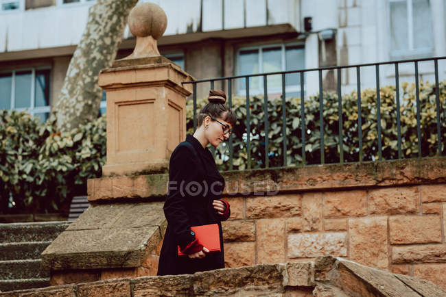 Giovane donna elegante in occhiali che tiene libro e scende sulle scale di fronte all'edificio in città — Foto stock