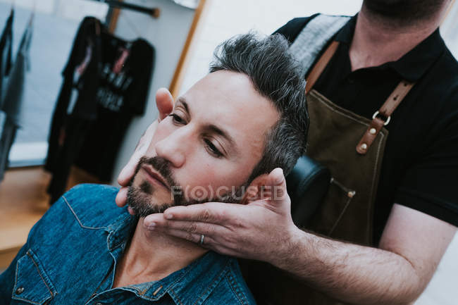 Barbiere facendo massaggio viso al bel maschio elegante con gli occhi chiusi seduti sulla sedia — Foto stock
