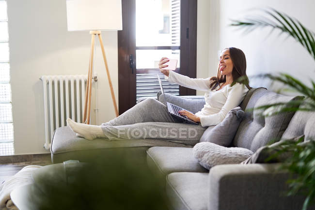Весела молода жінка, використовуючи ноутбук і приймаючи селфі з мобільним телефоном на дивані вдома — стокове фото