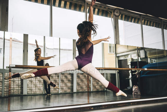 Vista trasera de la joven bailarina delgada saltando por encima del suelo en el estudio flexionando las piernas. - foto de stock