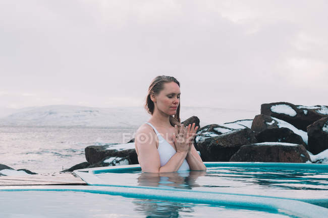 Mujer joven con los ojos cerrados meditando en el agua de la piscina cerca de rocas y cielo nublado - foto de stock