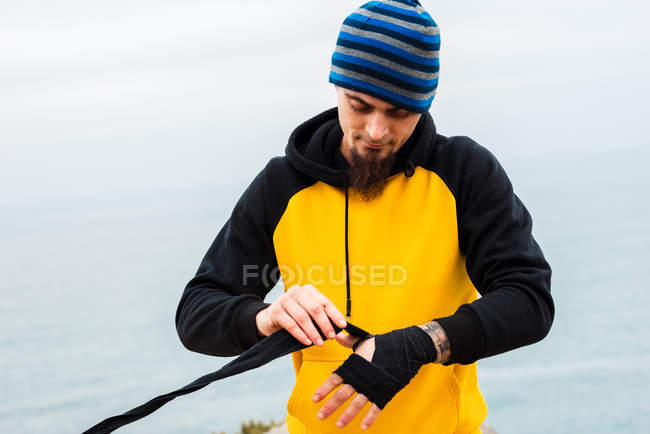 Бородатий дорослий чоловік обмотує пов'язку навколо руки, стоячи проти моря — стокове фото