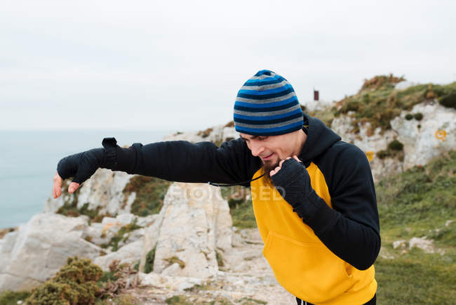 Homem barbudo adulto em sportswear praticando socos durante o treino de kickboxing em penhasco rochoso perto do mar — Fotografia de Stock