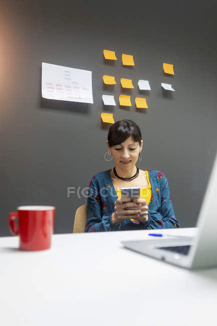 Mulher sorridente com caneca de bebida quente usando smartphone perto do laptop enquanto trabalhava no escritório moderno — Fotografia de Stock
