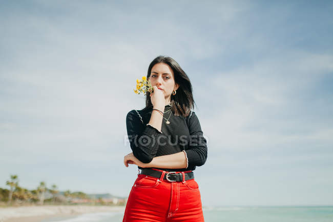 Чуттєва молода жінка з купою жовтих квітів, що стоять біля моря в сонячний день — стокове фото