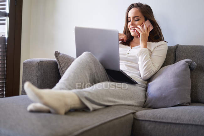 Souriant jeune femme heureuse à l'aide d'un ordinateur portable et parler sur un téléphone mobile sur le canapé à la maison — Photo de stock