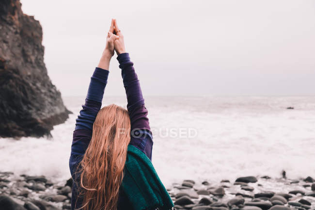 Young woman with upped hands standing on sea coast with pebbles in overcast — Stock Photo