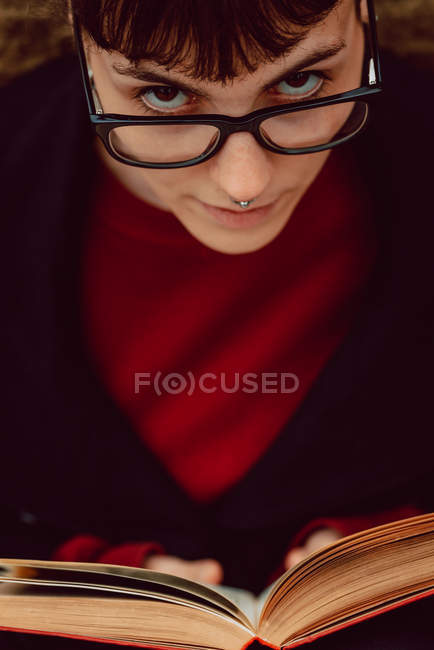 Nahaufnahme einer jungen eleganten Frau mit Brille und Buch, die in die Kamera blickt — Stockfoto