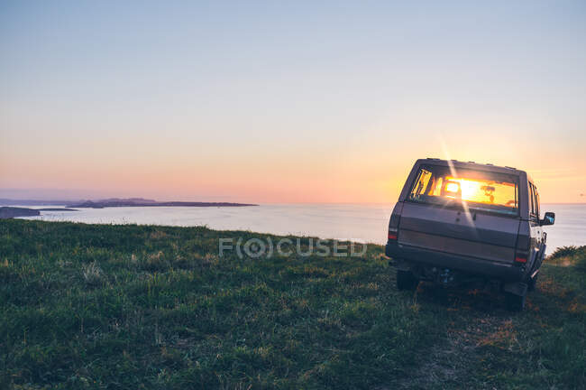 Moderno coche aparcado en la costa cubierta de hierba contra el mar tranquilo y el hermoso cielo al atardecer en Cantabria, España - foto de stock