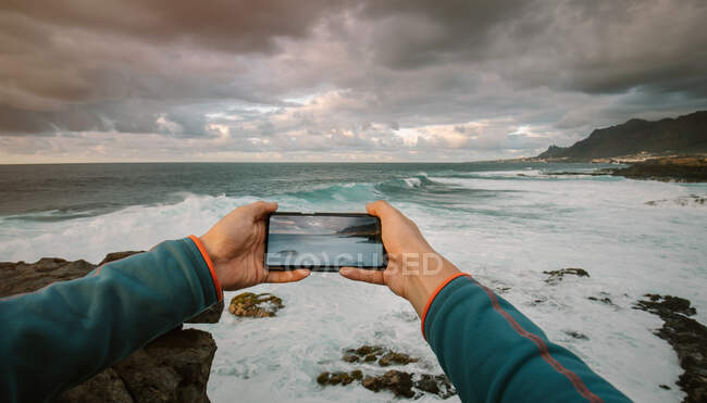 Vista en primera persona de un hombre tomando una foto con teléfono en un paisaje costero - foto de stock