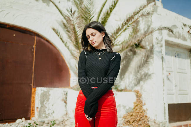 Young woman in trendy outfit with closed eyes standing near tropical palm leaves and white house on street — Stock Photo