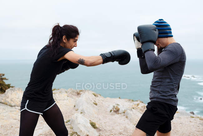 Hombre y mujer en guantes de boxeo golpeándose entre sí mientras están de pie en el acantilado contra el mar y el cielo - foto de stock
