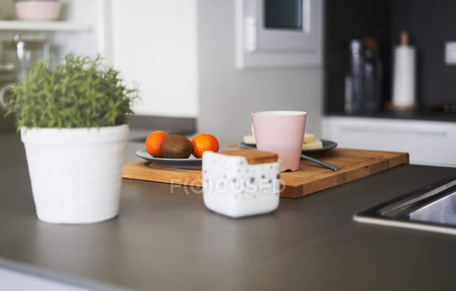Frutas em prato e xícara em tábua de corte de madeira e vaso de plantas na cozinha moderna — Fotografia de Stock