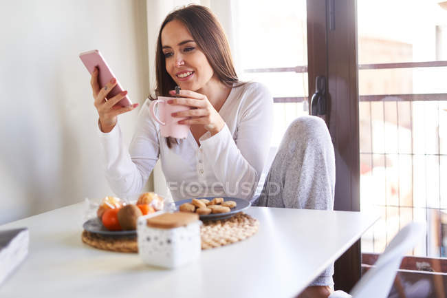 Jovem mulher feliz usando telefone celular e tomando café da manhã na mesa perto da janela em casa — Fotografia de Stock