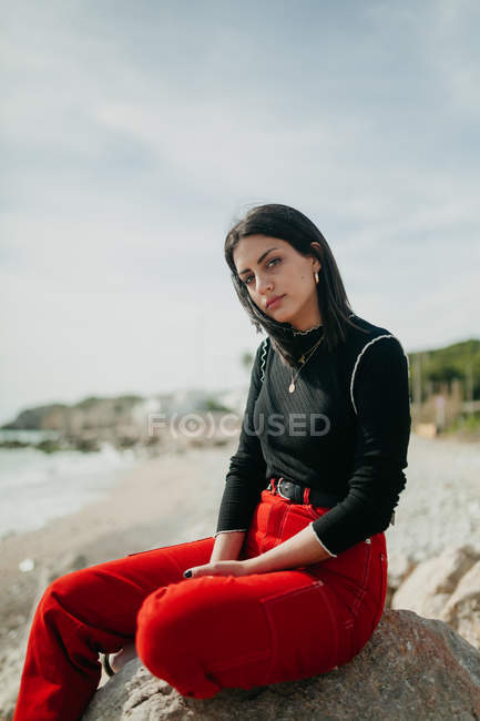 Mulher elegante olhando para a câmera na praia enquanto sentado na rocha no dia ensolarado — Fotografia de Stock