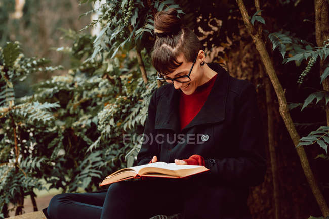 Joven mujer alegre elegante en anteojos libro de lectura mientras está sentado en el banco en el jardín de la ciudad - foto de stock