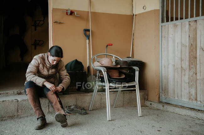 Молода жінка одягає захисний одяг, сидячи в стайні перед верхи на ранчо — стокове фото