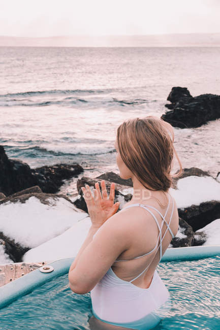 Junge Frau meditiert im Wasser des Pools in der Nähe von Felsen und bewölktem Himmel — Stockfoto