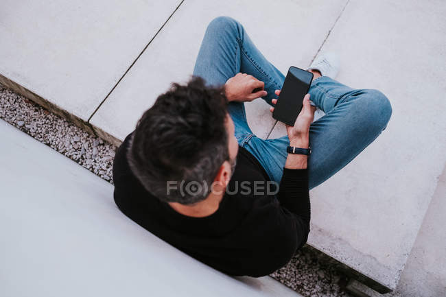 Idade Média bonito elegante macho feliz em uso casual usando um telefone celular e sentado perto da parede cinza — Fotografia de Stock