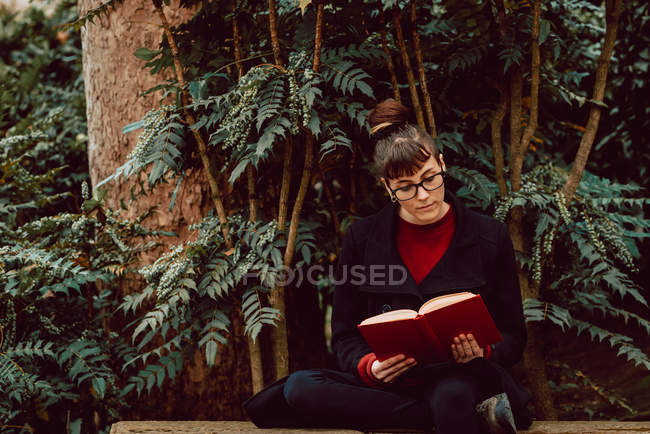 Giovane donna elegante in occhiali libro di lettura e seduta sulla panchina in giardino della città — Foto stock
