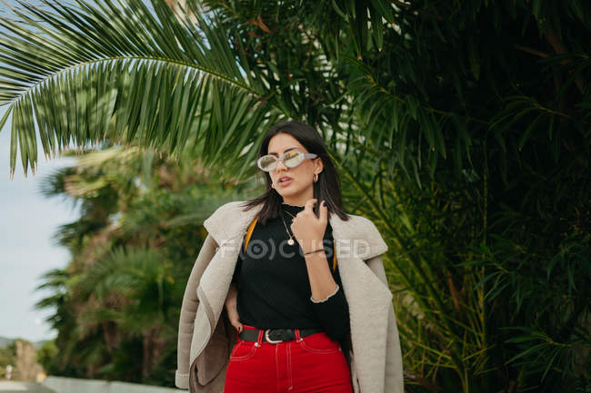 Молодая женщина в модном наряде позирует возле тропических пальмовых листьев на улице — стоковое фото