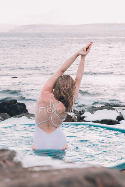 Visão traseira da mulher loira com as mãos levantadas relaxando na água da piscina perto de rochas em nublado — Fotografia de Stock