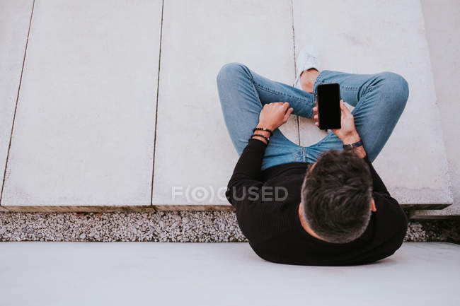 Edad media guapo elegante hombre feliz en ropa casual usando un teléfono móvil y sentado cerca de la pared gris - foto de stock