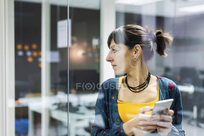 Gerente femenino sosteniendo teléfono inteligente mientras mira hacia otro lado cerca de la pared de vidrio en la oficina moderna - foto de stock