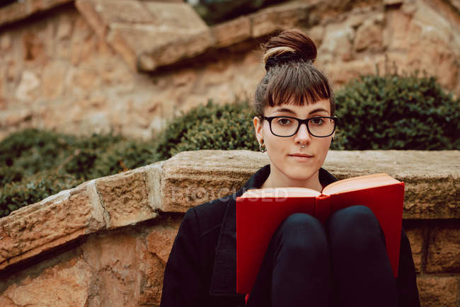 Junge elegante Frau mit Brille lehnt mit Buch an Steinmauer und blickt in die Kamera — Stockfoto