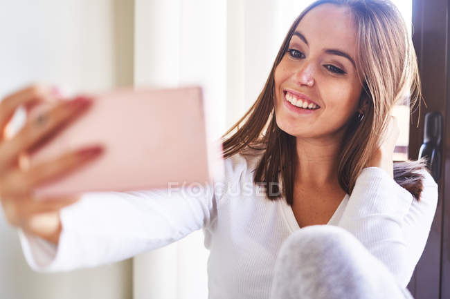 Jovem mulher feliz tomando selfie com telefone celular perto da janela em casa — Fotografia de Stock