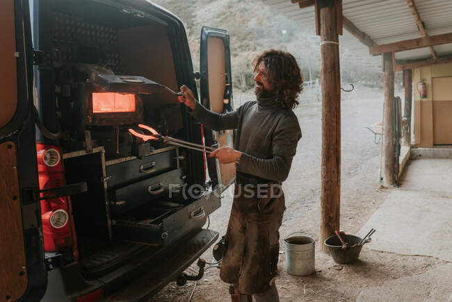 Bärtiger Hufschmied nimmt heißes Hufeisen aus tragbarem Ofen im Auto, während er in der Nähe des Stalls auf der Ranch arbeitet — Stockfoto