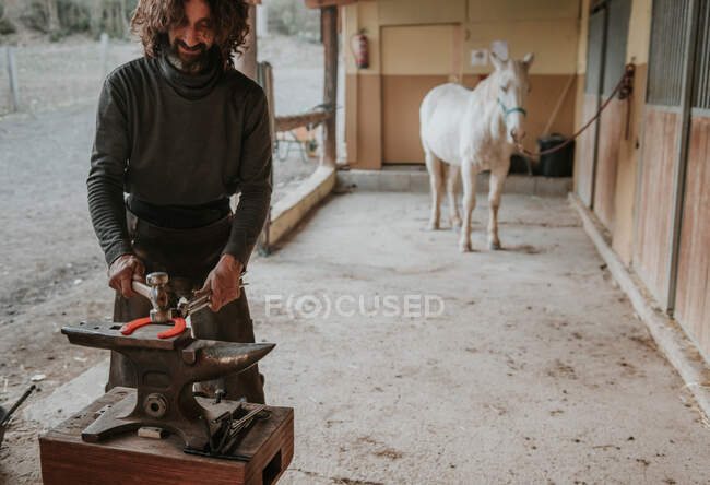 Взрослый кузнец, использующий молоток и щипцы для ковки горячей подковы на переносном наковальне возле конюшни на ранчо — стоковое фото