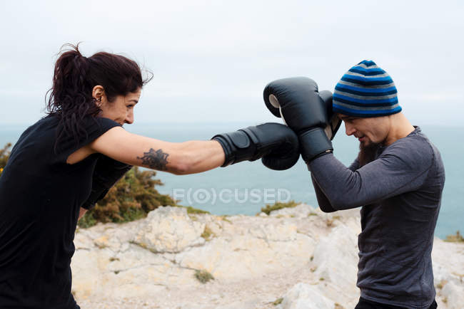 Чоловік і жінка в боксерських рукавичках вдарили один одного, стоячи на скелі проти моря і неба — стокове фото