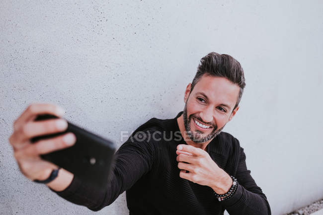 Gutaussehender eleganter, glücklicher Mann mittleren Alters in Freizeitkleidung, der ein Selfie mit dem Handy macht und in der Nähe einer grauen Wand sitzt — Stockfoto
